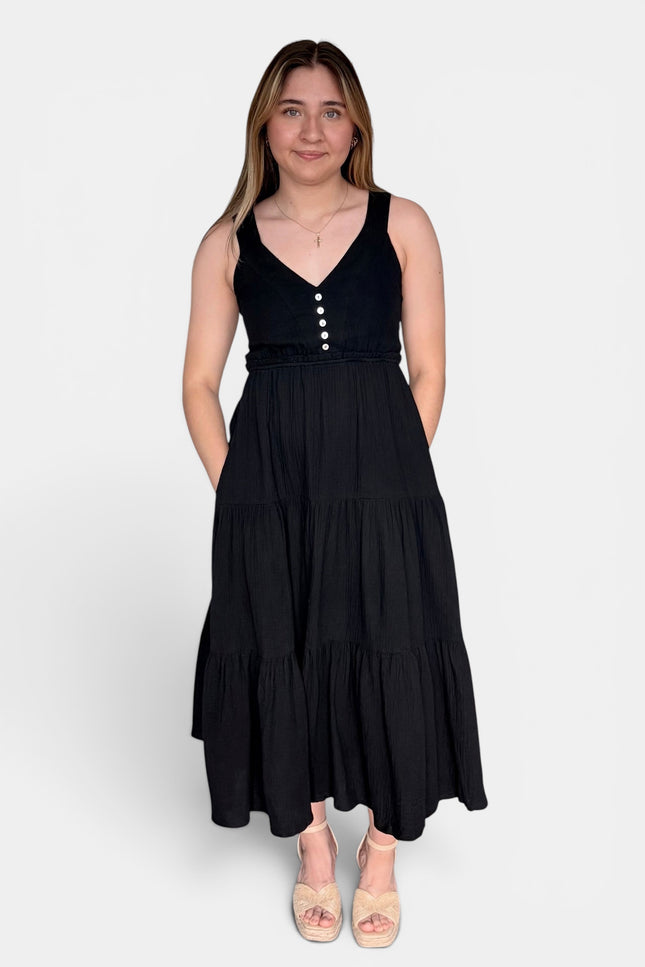 Black sleeveless tiered maxi dress with a V-neckline and decorative buttons down the bodice, paired with beige platform sandals, against a light gray studio background.