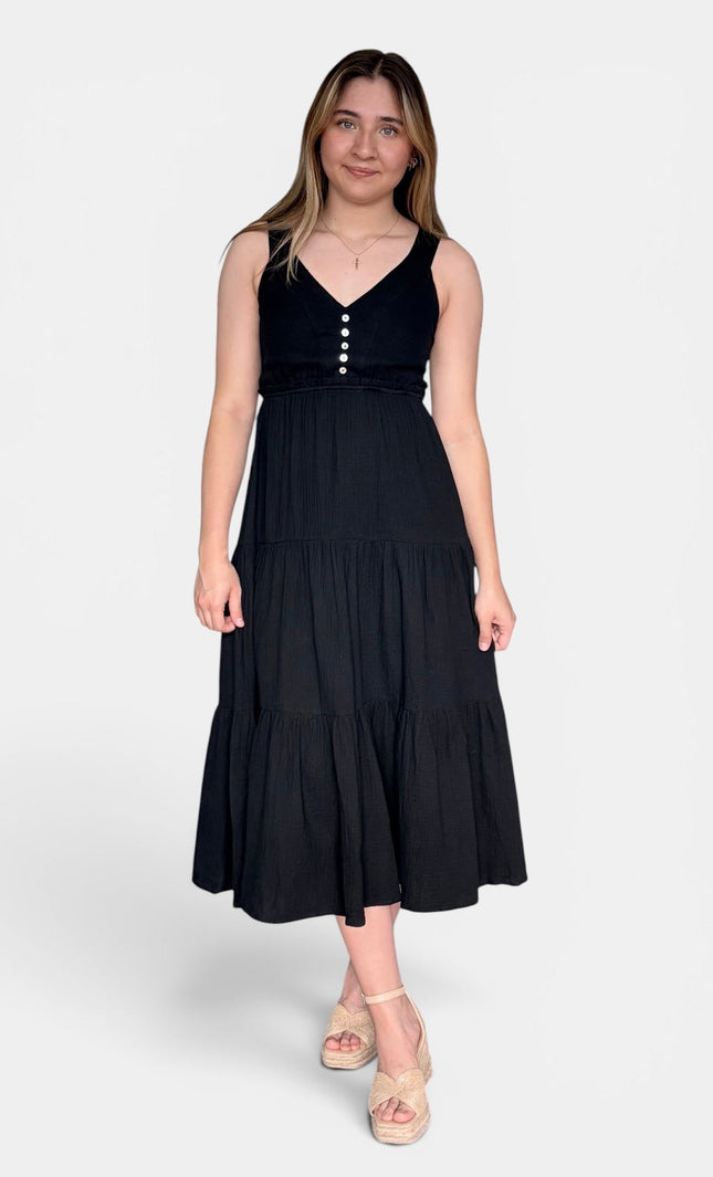 Sleeveless black midi dress with a V-neckline, buttoned bodice, and tiered skirt, paired with beige platform sandals against a light gray studio background.