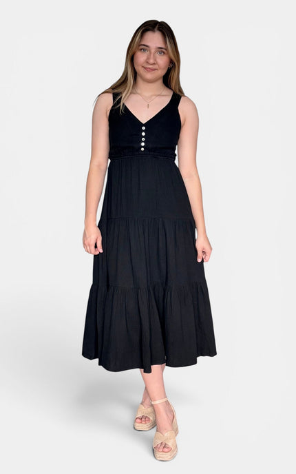 Sleeveless black midi dress with a V-neckline, buttoned bodice, and tiered skirt, paired with beige platform sandals against a light gray studio background.