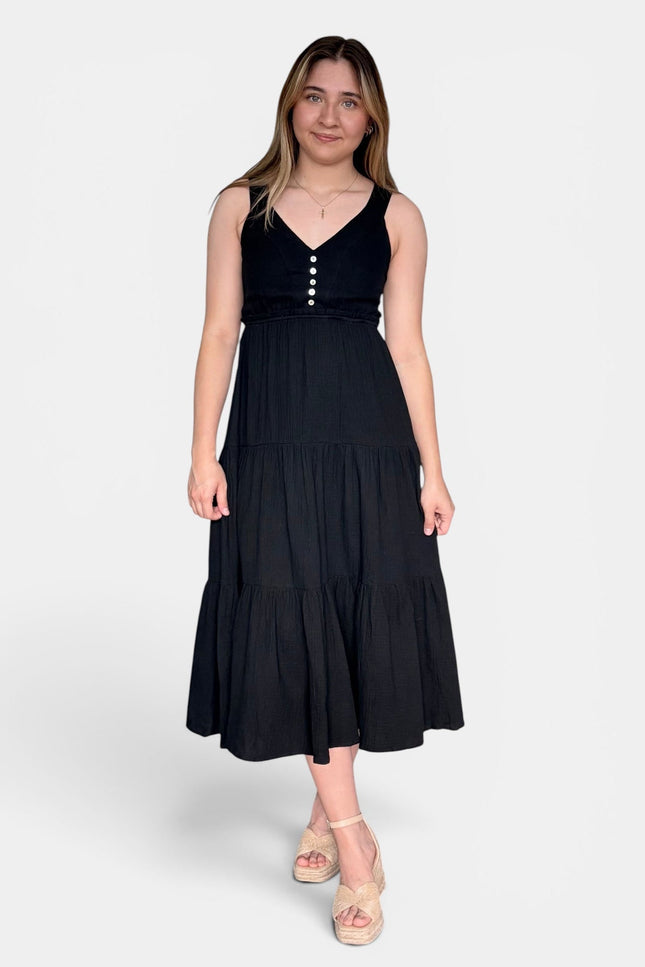 Sleeveless black midi dress with a V-neckline, buttoned bodice, and tiered skirt, paired with beige platform sandals against a light gray studio background.