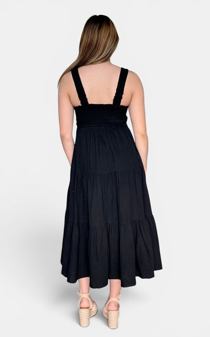Back of black sleeveless midi dress with shoulder straps and a tiered skirt, paired with beige platform sandals, against a light gray studio background.