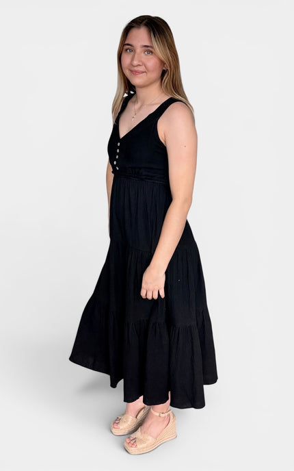 Sleeveless black midi dress with a tiered skirt and decorative buttons on the bodice, paired with beige flat sandals, against a light gray studio background.