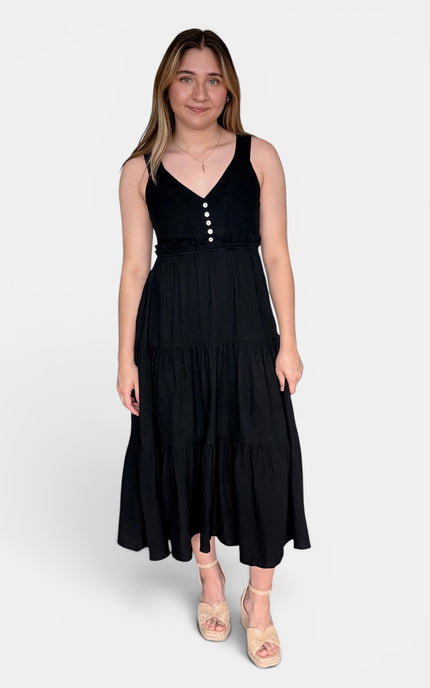 Sleeveless black midi dress with a tiered skirt and a buttoned V-neck bodice, paired with beige platform sandals, against a light gray studio background.