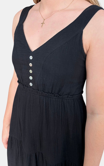 A close-up of a woman wearing a sleeveless black V-neck dress with a button-front bodice and gathered waist, against a plain light background.