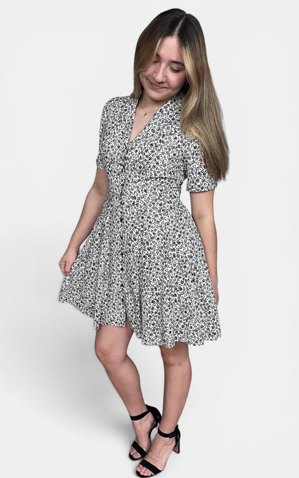 A woman modeling a short-sleeve button-front dress with a black and white floral print, paired with black strappy heels, against a light gray studio background.
