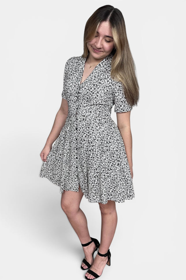 A woman modeling a short-sleeve button-front dress with a black and white floral print, paired with black strappy heels, against a light gray studio background.