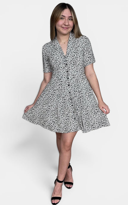Short-sleeve button-front dress with a black and white floral print, paired with black strappy heels, against a plain light gray studio background.
