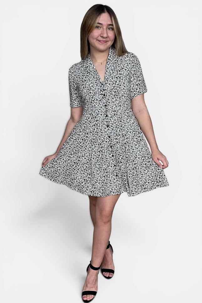 Short-sleeve button-front dress with a black and white floral print, paired with black strappy heels, against a plain light gray studio background.