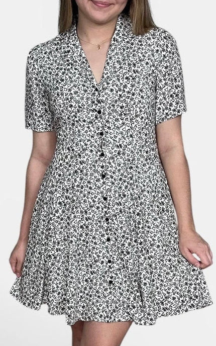Short-sleeve button-front dress with a black and white floral pattern, paired with black strappy heels, against a plain light gray studio background.