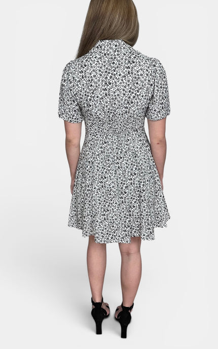A woman viewed from the back wearing a short-sleeve collared dress with an allover black and white floral print, paired with black high-heeled sandals, against a light gray studio background.