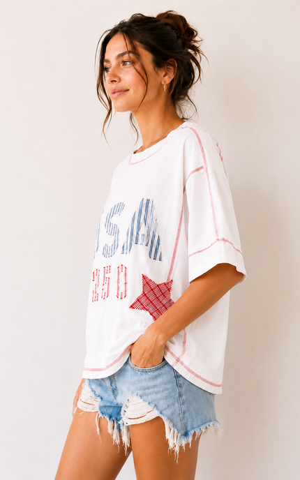 Woman wearing a white oversized t-shirt with text and graphics, paired with denim shorts, against a plain background.