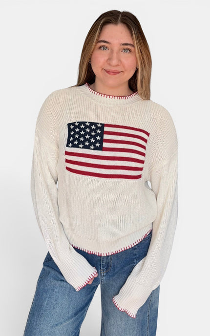 A woman wearing a cream-colored ribbed knit sweater with a red, white, and blue American flag graphic on the chest, paired with blue jeans, against a plain white background.