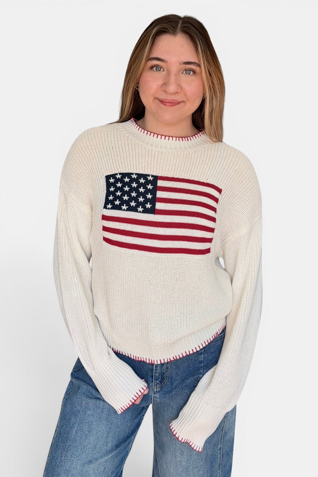 A woman wearing a cream-colored ribbed knit sweater with a red, white, and blue American flag graphic on the chest, paired with blue jeans, against a plain white background.