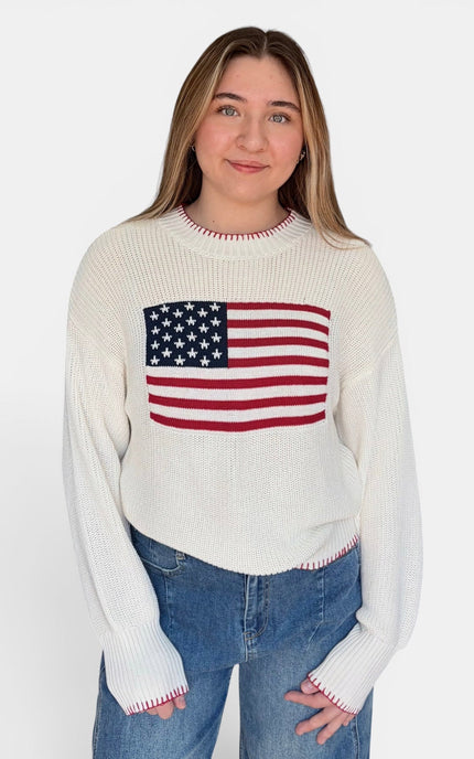 A woman wearing a cream-colored ribbed knit sweater with a red, white, and blue American flag graphic on the chest, paired with blue jeans, against a plain white studio background.