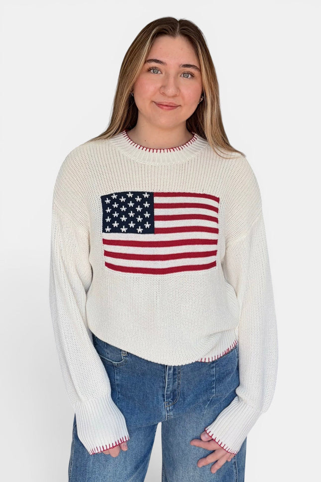 A woman wearing a cream-colored ribbed knit sweater with a red, white, and blue American flag graphic on the chest, paired with blue jeans, against a plain white studio background.