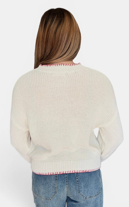 A woman viewed from the back wearing a cream-colored knit sweater with red striped trim at the collar and hem, paired with blue jeans, against a plain white background.