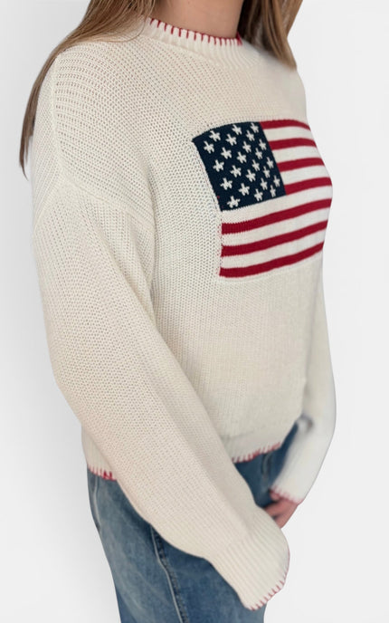 A woman wearing a cream-colored knit sweater with a large American flag graphic on the chest, paired with blue jeans, shown against a plain light gray background.