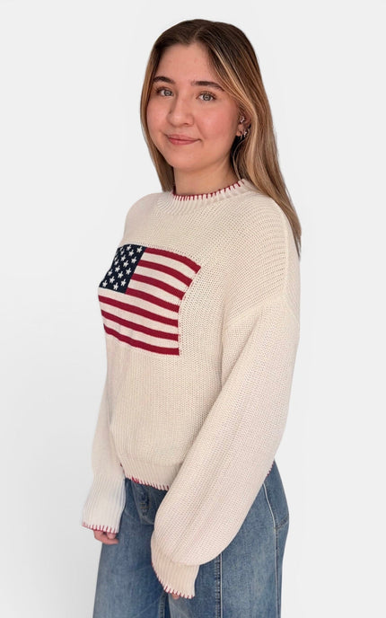 A woman wearing a cream-colored knit sweater with a red, white, and blue American flag graphic on the chest, paired with blue jeans, photographed against a plain white background.
