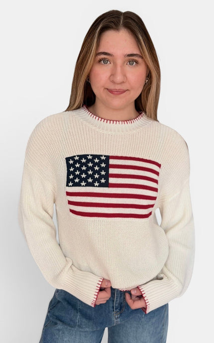 A young woman wearing a cream-colored knit sweater with a large American flag graphic on the chest, paired with blue jeans, against a plain light gray background.