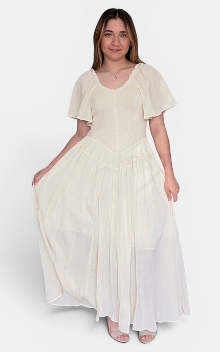 A woman modeling a cream-colored maxi dress with a textured bodice, flutter sleeves, and a flowing skirt, photographed against a plain light gray studio background.