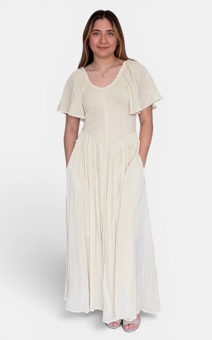A woman modeling a cream-colored maxi dress with short flutter sleeves, a textured bodice, and a flowing skirt, standing against a light gray studio background.