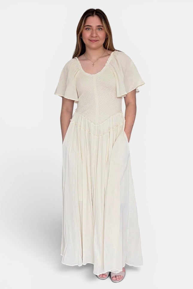 A woman modeling a cream-colored maxi dress with short flutter sleeves, a textured bodice, and a flowing skirt, standing against a light gray studio background.