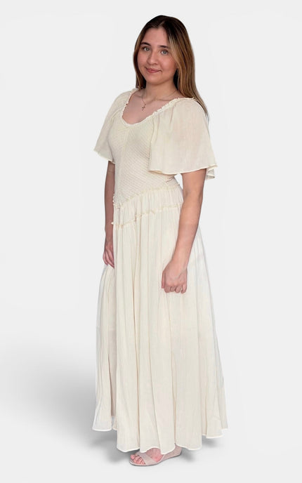 A woman modeling a cream-colored maxi dress with short flutter sleeves, a scoop neckline, and a tiered skirt, photographed against a light gray studio background.