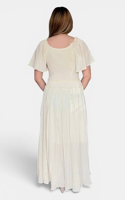 A woman viewed from the back wearing a long cream-colored dress with short flutter sleeves, a smocked bodice, and a flowing skirt, photographed against a light gray studio background.