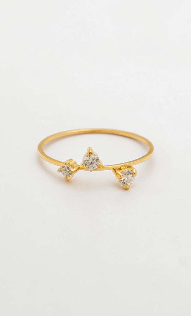 A minimalist flat lay of a delicate 18k gold three-stone CZ ring resting on a clean, soft background, showcasing its timeless and romantic aesthetic.