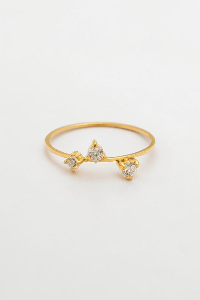 A minimalist flat lay of a delicate 18k gold three-stone CZ ring resting on a clean, soft background, showcasing its timeless and romantic aesthetic.