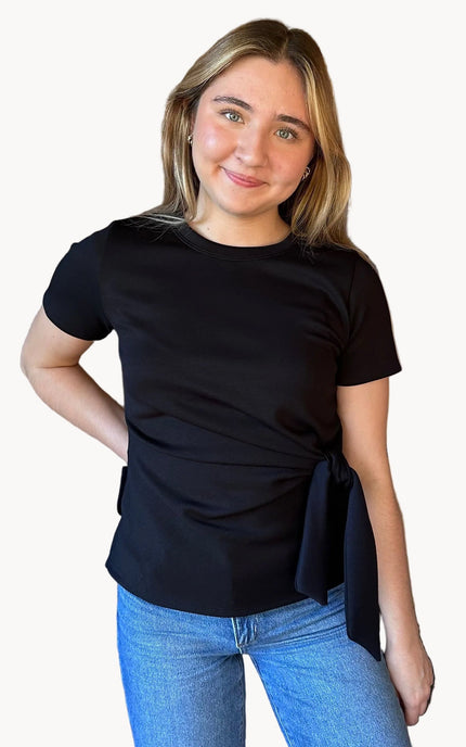 Woman wearing a black top with a tie detail and blue jeans on a beige background