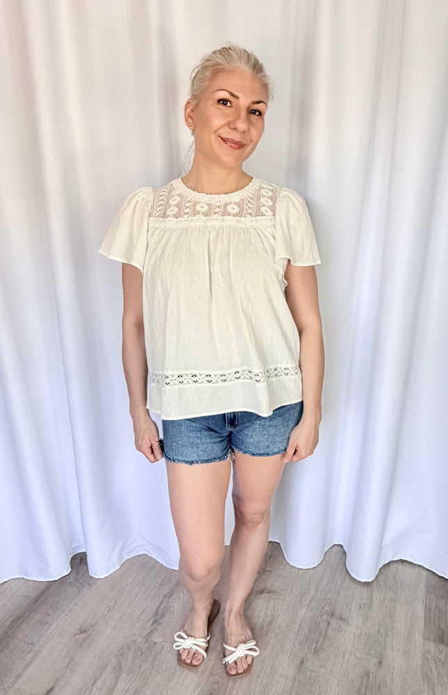 Front view of a model wearing the white cotton Aurelia blouse tucked loosely into high-waisted bottoms, highlighting the flowy drape of the bodice and the delicate placement of the lace yoke.