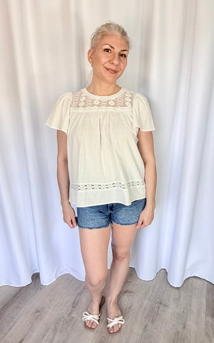 Front view of a model wearing the white cotton Aurelia blouse tucked loosely into high-waisted bottoms, highlighting the flowy drape of the bodice and the delicate placement of the lace yoke.