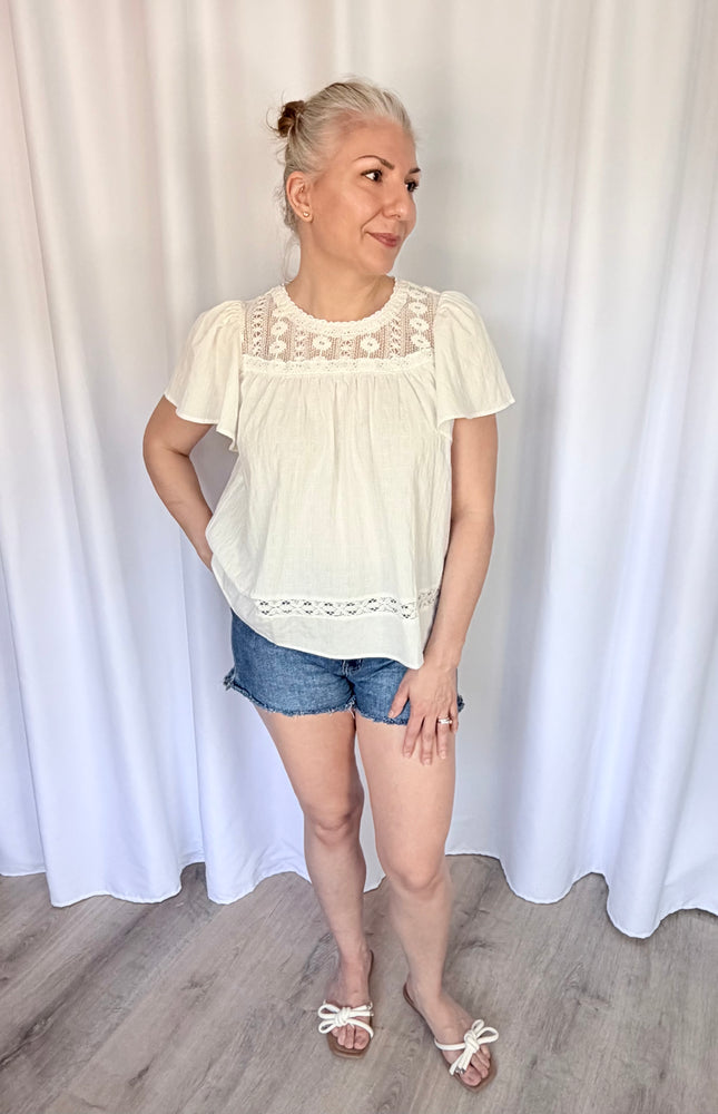 A woman wearing the white cotton Aurelia blouse, showcasing its relaxed, flowy silhouette and short flutter sleeves. The top features a round neckline and intricate, vintage-inspired lace detailing across the chest yoke.