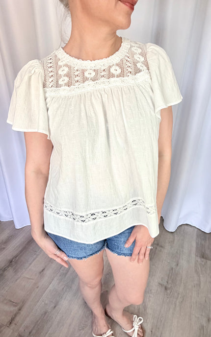 A waist-up front view of the Aurelia blouse, focusing on the romantic combination of the round neckline, intricate vintage lace details, and the soft flare of the short flutter sleeves.