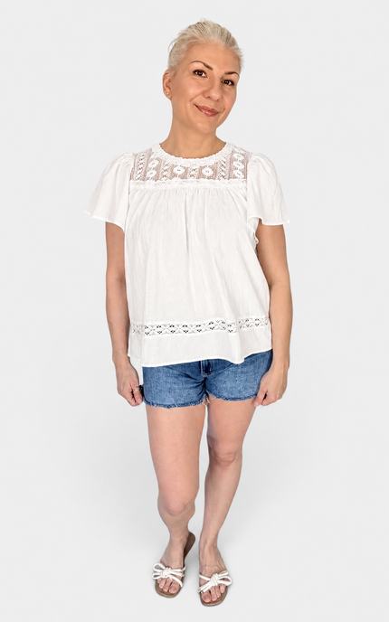 A woman modeling a white short-sleeve blouse with eyelet lace trim at the neckline and hem, paired with denim shorts and white sandals, against a light gray studio background.