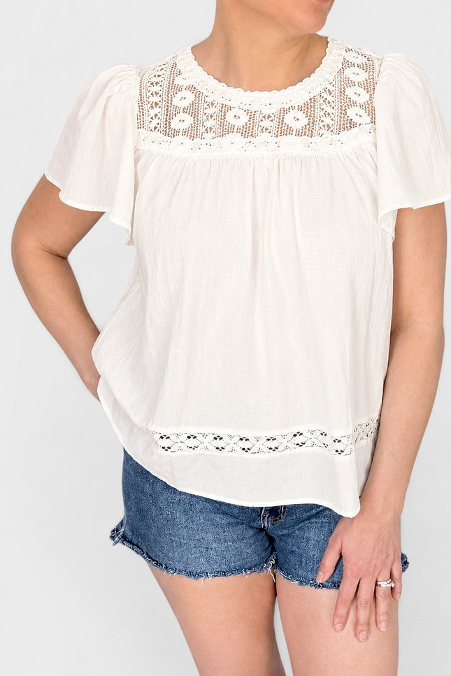 A woman wearing a white short-sleeve blouse with delicate lace trim at the round neckline and hem, paired with denim shorts, against a light gray studio background.