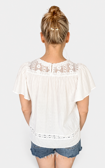 A woman viewed from the back wearing a white short-sleeve blouse with lace trim at the neckline and hem, paired with denim shorts, against a light gray studio background.