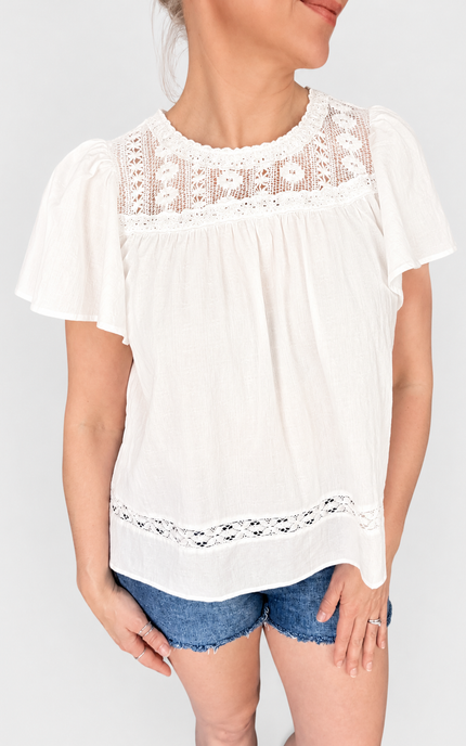 A woman wearing a white short-sleeve blouse with delicate lace trim at the neckline and hem, paired with denim shorts, against a light gray studio background.