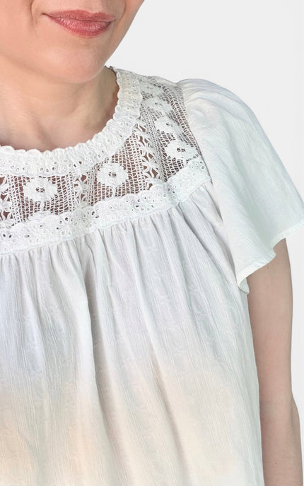 A close-up of a woman wearing a white short-sleeve blouse with a delicate floral lace yoke and gathered neckline, set against a plain light background.
