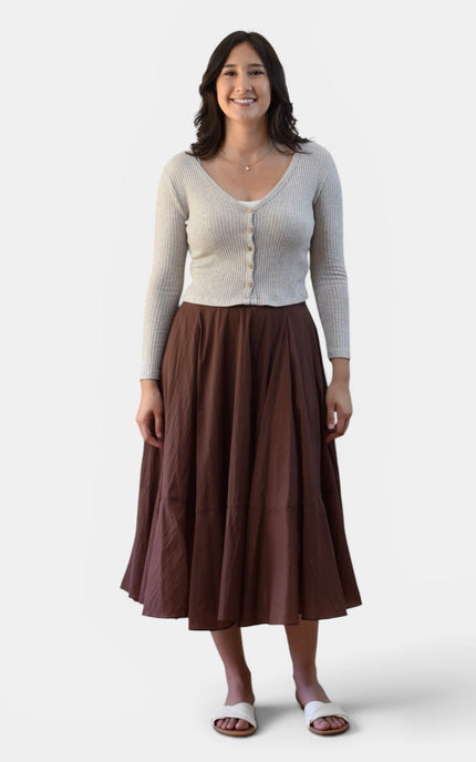A woman modeling a light beige ribbed cardigan with buttons over a brown tiered midi skirt, paired with white slide sandals, against a plain light gray background.
