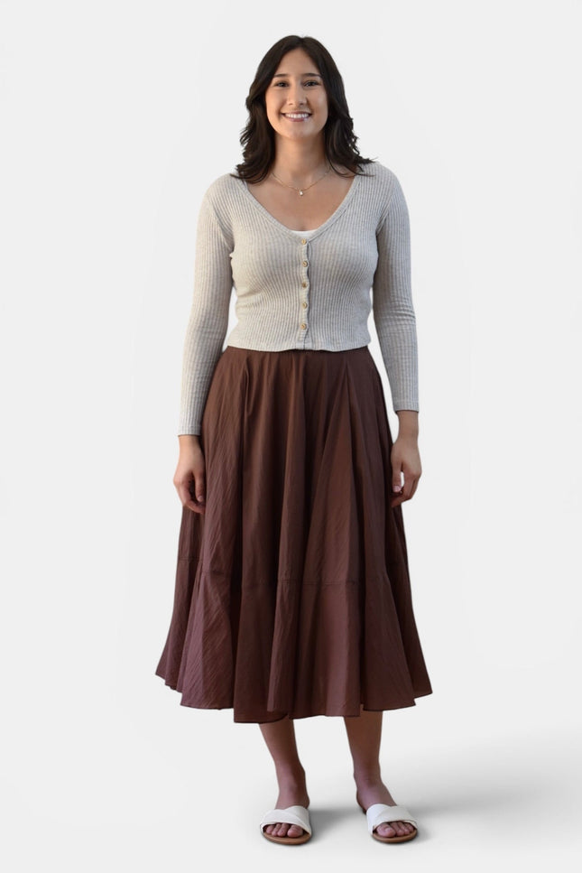 A woman modeling a light beige ribbed cardigan with buttons over a brown tiered midi skirt, paired with white slide sandals, against a plain light gray background.
