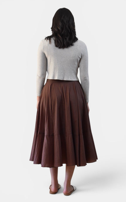 A woman shown from the back wearing a light gray ribbed knit long-sleeve top and a brown pleated midi skirt, standing against a plain light gray studio background.
