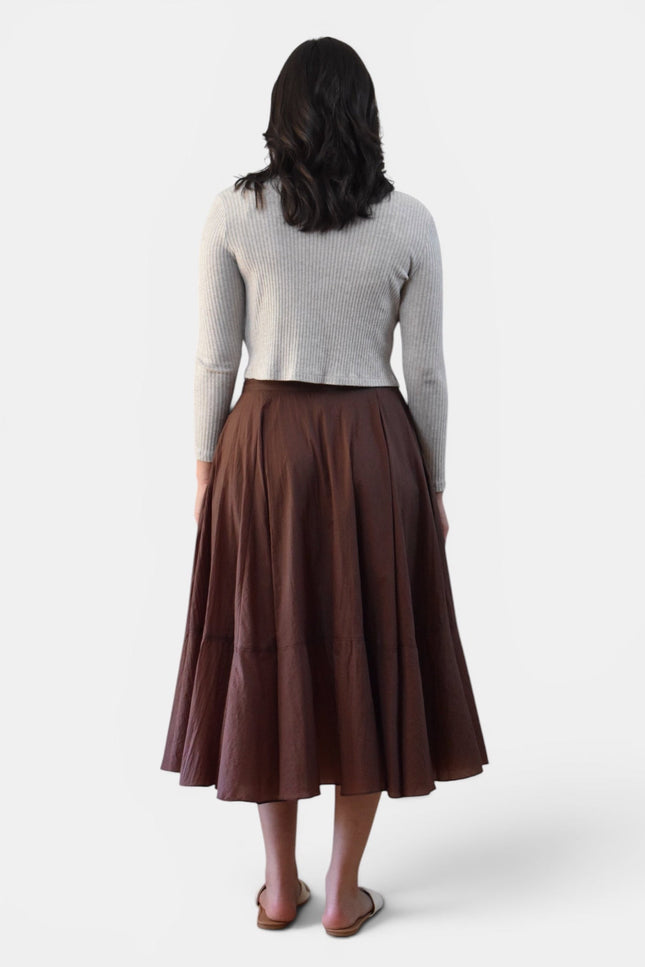 A woman shown from the back wearing a light gray ribbed knit long-sleeve top and a brown pleated midi skirt, standing against a plain light gray studio background.