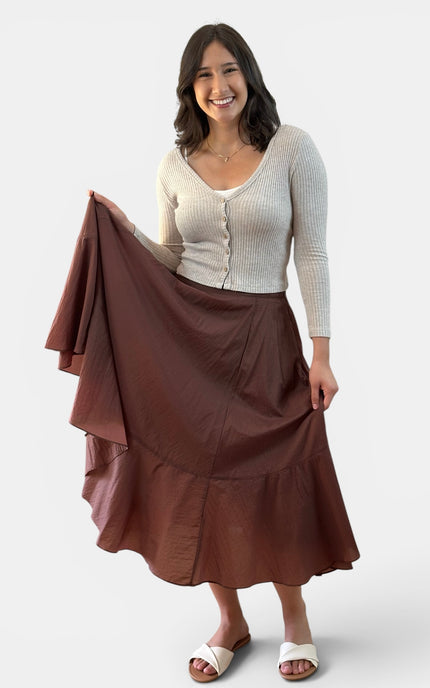 A woman modeling a brown tiered midi skirt with a matching fabric drape, paired with a cream ribbed cardigan and white slide sandals, against a light gray studio background.
