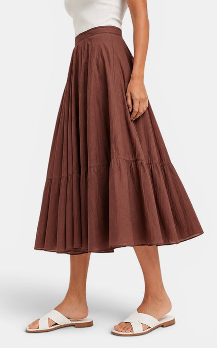 A model wearing a brown tiered midi skirt with a white top and white slide sandals, shown from the waist down against a light gray studio background.