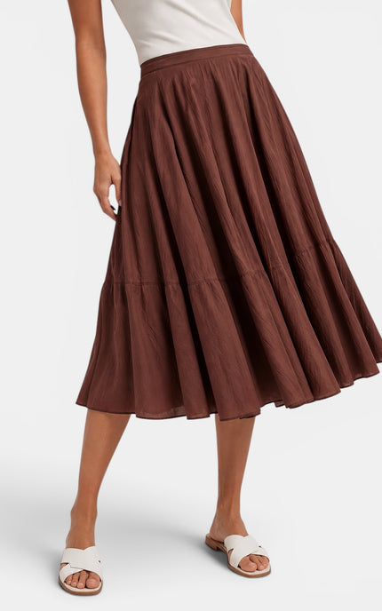 A brown tiered midi skirt worn by a model, paired with a white top and white slide sandals, shown against a light gray studio background.