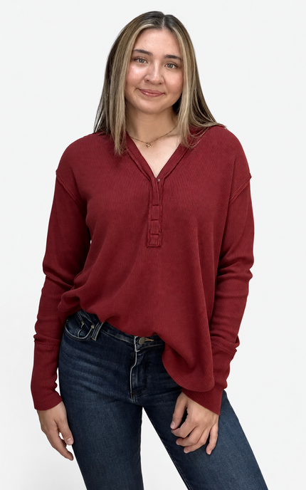 A woman wearing a long-sleeve burgundy henley top and dark blue jeans, standing against a plain light gray background.
