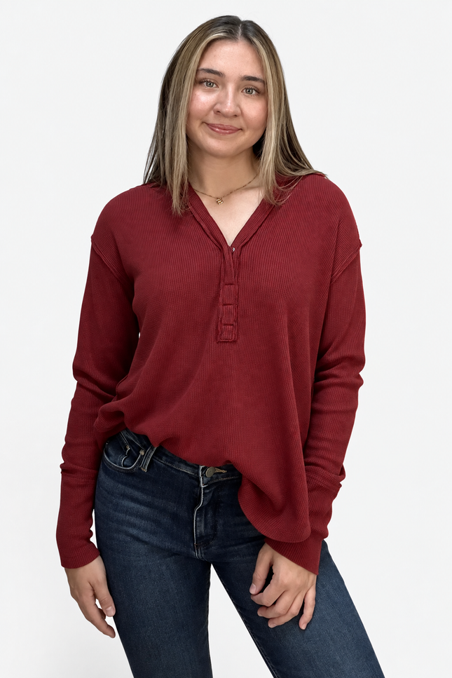 A woman wearing a long-sleeve burgundy henley top and dark blue jeans, standing against a plain light gray background.