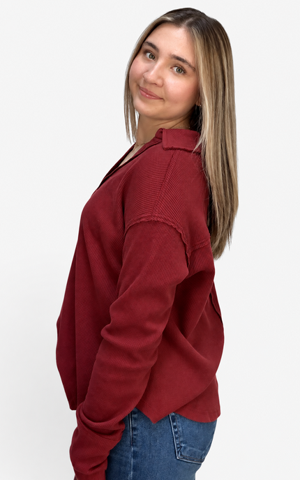 A woman with blonde-highlighted hair wearing a dark red knit button-up shirt and blue jeans, photographed from the side against a plain white studio background.